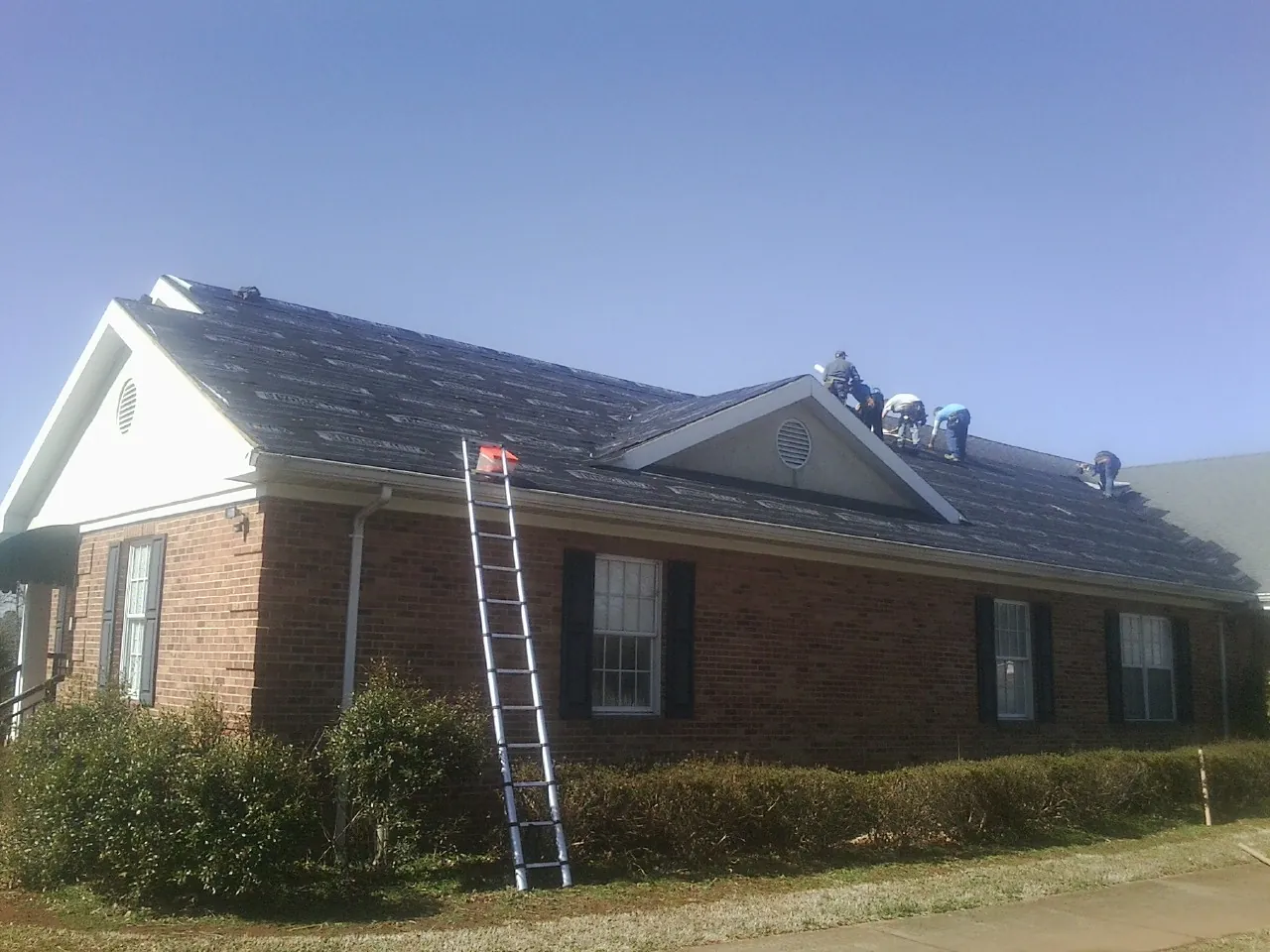 Crew tearing shingles off a brick ranch roof with a ladder set — Emergency Roof Repair work by our crew in Bentonia