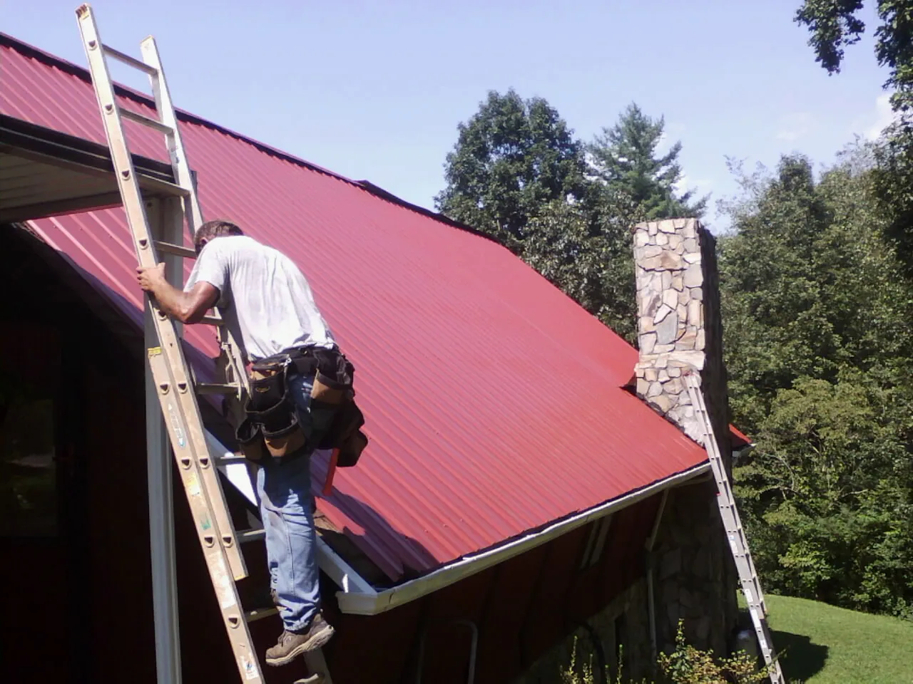 Roofer on a ladder beside a red metal roof and stone chimney — Soffit & Fascia Repair by Roofing Brothers in Bentonia